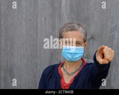 Die kaukasische Frau mittleren Alters trägt eine blaue chirurgische Gesichtsmaske und zeigt mit ihrem Zeigefinger auf den Betrachter während der Corona-Pandemie von 2019/20. Stockfoto