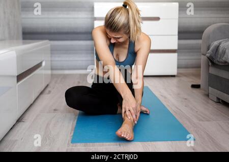 Unglückliche Frau, die auf der Yogamatte sitzt, mit Knöchelverletzung, Schmerz fühlend. Gesundheitswesen und execise Konzept. Stockfoto