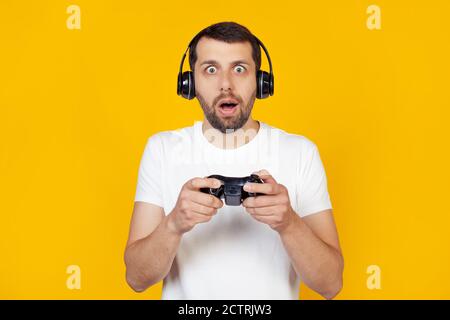 Junger Mann mit Bart in weißem T-Shirt Gamer spielt Videospiele mit Kopfhörern, erschrocken im Schock mit überrascht Gesicht, erschrocken und aufgeregt mit Angst Ausdruck steht auf isolierten gelben Hintergrund. Stockfoto
