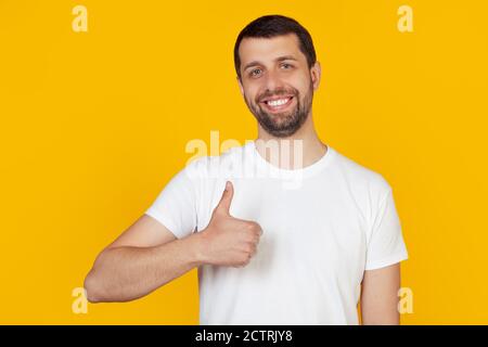 Junger Mann mit einem Bart in einem weißen T-Shirt macht eine glückliche Hand Geste. Bestätigung des Gesichtsausdrucks, der Erfolg beim Betragen der Kamera zeigt. Steht auf isoliertem gelben Hintergrund. Stockfoto