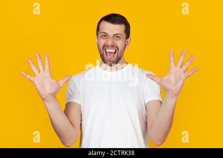 Ein junger Mann mit einem Bart in einem weißen T-Shirt ist sehr glücklich und aufgeregt, macht eine Gewinnergeste mit erhobenen Händen, lächelt und schreit Erfolg. Celebration Concept steht auf isoliertem gelben Hintergrund Stockfoto