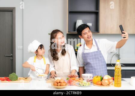 Junge asiatische Familie bereiten Salat in der Küche und Vater nehmen ein Foto Selfie per Telefon. Aufgeregt lächeln und Fällen glücklich. Eltern lehren Tochter Stockfoto