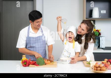 Asiatische Familie bricht ein Ei in die Schüssel und Tochter Rühreier in Schüssel auf weißem Tisch und sieht glücklich aus Essen kochen mit ihrem Vater und mot Stockfoto