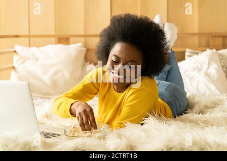 Fröhliche Afro-Amerikanerin in gelbem Pullover, die sich entspannt, Popcorn isst, während sie sich einen Film auf dem Laptop ansieht, im Video-Chat spricht, im Bett liegt. Faule Ruhe an Stockfoto