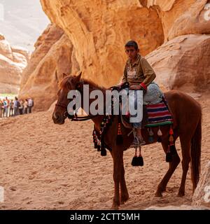 Petra, Jordanien 04/02/2010: Ein beduinenjunge aus der Region trägt Jeans und Sandalen und ist auf einem Pferd mit dekorativem Mundgebiss. Sie ruhen im Schatten einer Klippe in Stockfoto