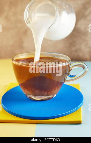 Eine Tasse Kaffee und Milch fließt in eine Tasse auf einem gelben und blauen Podium. Stockfoto