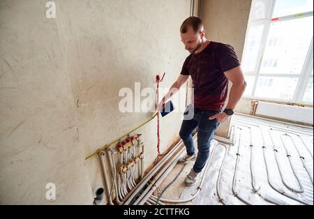 Serious Mann Messung Wand mit Band Maß im Raum mit Fußbodenheizung Rohre. Handyman hält Notebook und Messungen während der Arbeit an der Renovierung der Wohnung. Reparaturkonzept