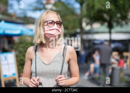 Porträt von Casual junge Frau auf der Straße zu Fuß in Schutzmaske als Schutz gegen covid-19-Virus. Beiläufige Personen im Hintergrund Stockfoto