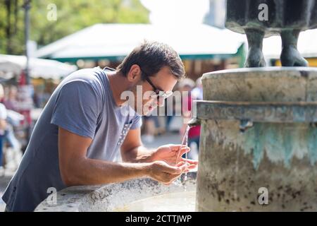 Durstige junge Casual cucasian Mann Trinkwasser aus der öffentlichen Stadt Brunnen an einem heißen Sommertag Stockfoto