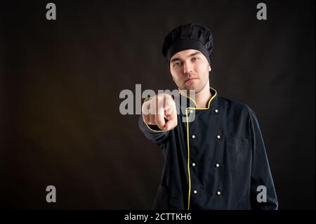 Junger Mann in einem schwarzen Koch Anzug gekleidet zeigt seine Finger posiert auf einem braunen Hintergrund mit Copy Space Werbung Bereich Stockfoto