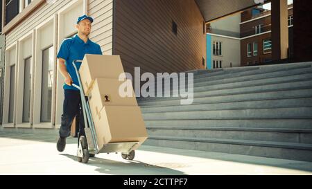 Happy Young Delivery man schiebt Hand Truck Trolley voll von Karton-Boxen und Pakete für die Lieferung. Professionelle Kurier arbeiten effizient und Stockfoto