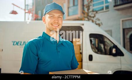 Portrait von Handsome Delivery man hält Karton-Box-Paket stehen in modernen stilvollen Business District mit Lieferwagen im Hintergrund. Lächelnd Stockfoto