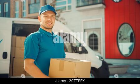 Portrait von Handsome Delivery man hält Karton-Box-Paket stehen in modernen stilvollen Business District mit Lieferwagen im Hintergrund. Lächelnd Stockfoto