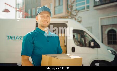 Portrait von Handsome Delivery man hält Karton-Box-Paket stehen in modernen stilvollen Business District mit Lieferwagen im Hintergrund. Lächelnd Stockfoto