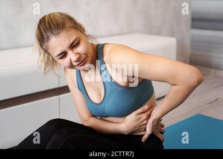 Porträt einer unglücklichen jungen Frau, die auf einer Yogamatte sitzt, nach dem Training den Rücken berührt, unter Rückenschmerzen leidet, Schmerzen spürt, Seitenansicht Stockfoto