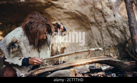 Ur Höhlenmensch trägt Tierhaut versucht, ein Feuer mit Bow Drill Methode zu machen. Neanderthal anzündet das erste menschengemachte Feuer in der menschlichen Zivilisation Stockfoto