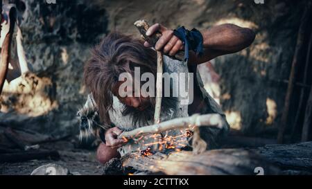 Nahaufnahme eines urzeitlichen Höhlenmenschen, der Tierhaut trägt und versucht, mit der Bow Drill Methode Feuer zu machen. Neanderthal anzündet das erste menschengemachte Feuer im Menschen Stockfoto