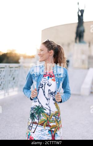 Junge fröhliche städtische Frau hält Denim Jacke an berühmten Ort Monument bei Sonnenuntergang Stockfoto