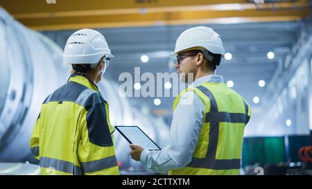 Zwei Schwerindustrie-Ingenieure stehen in der Pipe Manufacturing Factory, verwenden digitale Tablet-Computer, haben Diskussion. Bau von Öl, Gas und Brennstoffen Stockfoto