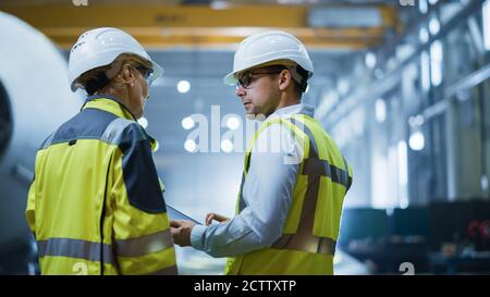 Zwei Schwerindustrie-Ingenieure stehen in der Pipe Manufacturing Factory, verwenden digitale Tablet-Computer, haben Diskussion. Bau von Öl, Gas und Brennstoffen Stockfoto