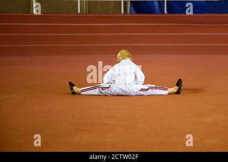 Ein Karate-Junge in einem weißen Kimono mit einem gelben Gürtel, der die Beine dehnt und dabei Garn auf dem Boden macht. Stockfoto