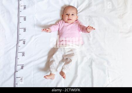 Ein viermonatiges glückliches Baby in rosa weißer Kleidung, das auf einem Bett liegt, auf dem ein Maßlineal für Wachstum gezeichnet wird. Stockfoto