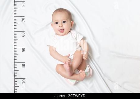 Ein fünfmonatiges Baby in weißer Kleidung, das auf einem Bett liegt, auf dem ein Maßlineal für Wachstum gezeichnet wird. Zeigt die Zunge aus ihrem Mund und gehänselt Stockfoto