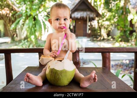 Ein trauriges, einjähriges Kind sitzt auf einem Holztisch und trinkt Kokosmilch aus frischer grüner Kokosnuss durch einen Strohhalm. Stockfoto