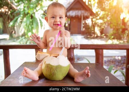 Ein fröhliches und glückliches, einjähriges Kind sitzt auf einem Holztisch und trinkt Kokosmilch aus frischer grüner Kokosnuss durch einen Strohhalm, den er in der Hand hält. Stockfoto