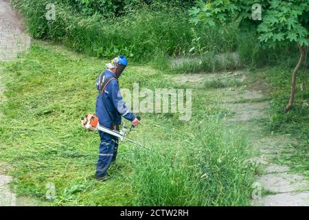 Mann in der Arbeit blauen Overalls mit einer Schutzmaske mäht das Gras mit Elektrowerkzeug, Handmäher, Schnur Rasenmäher Stockfoto