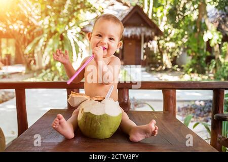 Ein fröhliches und glückliches, einjähriges Kind sitzt auf einem Holztisch und trinkt Kokosmilch aus frischer grüner Kokosnuss durch einen Strohhalm, den er in der Hand hält. Stockfoto