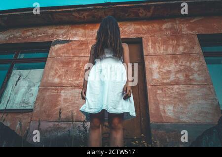 Mädchen in einem weißen Kleid mit langen dunklen Haaren über ihr Gesicht geworfen steht auf den Stufen eines verlassenen Gebäudes. Konzept des Grauens, Mystik Stockfoto