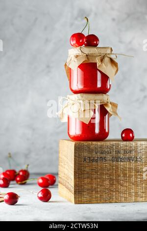 Hausgemachte Kirschmarmelade im Glas auf der Holzkiste auf dem grauen Hintergrund. Zwei Gläser Kirschkonfitüre auf Holz mit Kirschen im Rücken. Food-Fotografie. Stockfoto