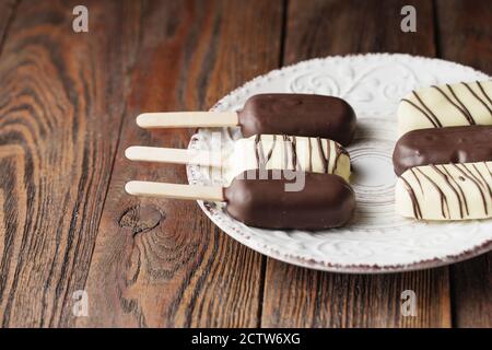Milchschokolade-Eis auf einem Stock. Eisdielen mit weißer und dunkler Schokolade auf dem Teller auf Holzhintergrund bedeckt. Leerzeichen für Text. Choc Stockfoto