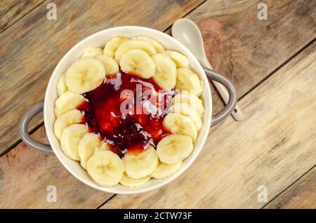 Haferflocken mit Marmelade und Bananen in einer weiß-grauen Platte mit einem weißen Löffel auf einem hölzernen Hintergrund Nahaufnahme, Draufsicht. Gesundes Frühstück. Stockfoto