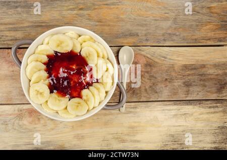 Haferflocken mit Marmelade und Bananen in einer weiß-grauen Platte mit einem weißen Löffel auf einem Holzhintergrund, Draufsicht, Kopierraum. Gesundes Frühstück. Stockfoto