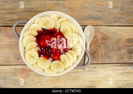 Haferflocken mit Marmelade und Bananen in einer weiß-grauen Platte mit einem weißen Löffel auf einem Holzhintergrund, Draufsicht, Kopierraum. Gesundes Frühstück. Stockfoto