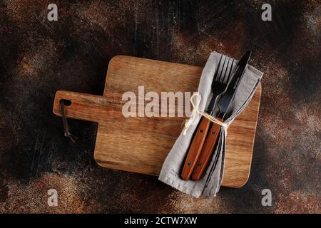 Besteck auf dem Teller auf dunkelbraunem Hintergrund. Gabel und Tischmesser mit Holzgriffen auf Leinenserviette und Holzdiele Stockfoto