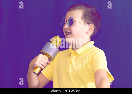 Nahaufnahme Bild.der junge Kind Kind 9s alt in gelben T-Shirt und blauen Brillen singt Musik auf lila neblig getönten beleuchteten Bühne. Stockfoto