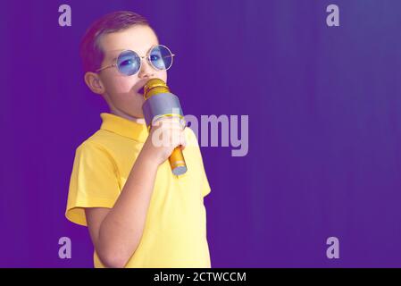 Nahaufnahme Bild.der junge Kind Kind 9s alt in gelben T-Shirt und blauen Brillen singt Musik auf lila neblig getönten beleuchteten Bühne. Stockfoto