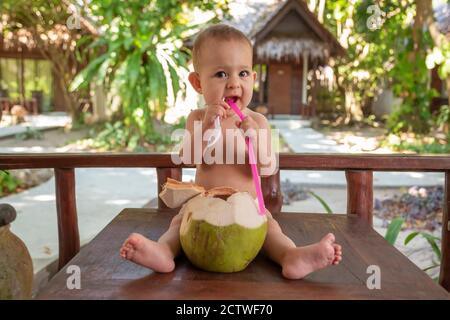 Ein fröhliches und glückliches, einjähriges Kind sitzt auf einem Holztisch und trinkt Kokosmilch aus frischer grüner Kokosnuss durch einen Strohhalm. Rechte Hand hält einen Löffel. Stockfoto