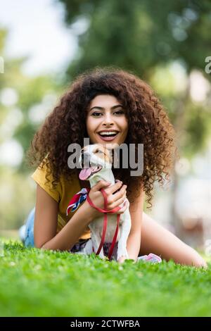 Aufgeregt Frau lachend und Blick auf die Kamera, während sie mit liegen jack russell Terrier Hund auf grünem Rasen Stockfoto