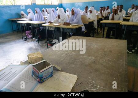 PEMBA ISLAND, SANSIBAR, TANSANIA - JANUAR 2020: Schulmädchen und Schuljungen im normalen Klassenraum mit einem Teachers Table mit Choke Box an Stockfoto