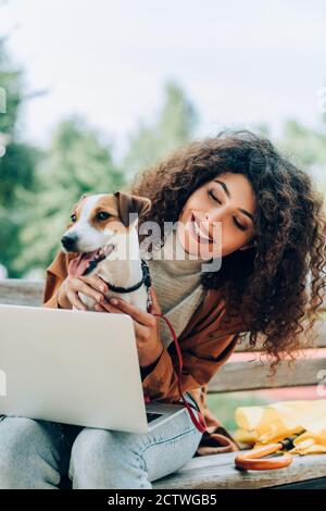 Fröhlicher Freiberufler kuscheln Jack russell Terrier Hund während des Sitzens auf Bank mit Laptop Stockfoto