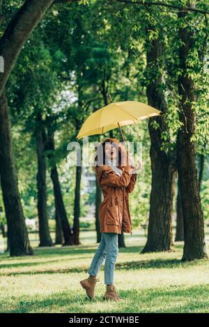 Junge Frau im Herbst Outfit mit gelben Regenschirm im Stehen Auf dem Rasen im Park Stockfoto