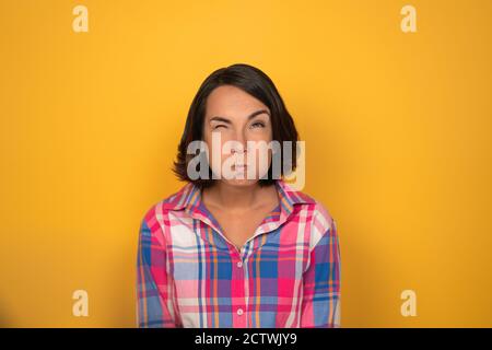 Die junge Frau hatte ihre Wangen auf einem gelben Hintergrund im Studio aufgepumpt. Sie hat ihre Augenbraue gebogen und schaut nach oben. Hochwertige Fotos Stockfoto