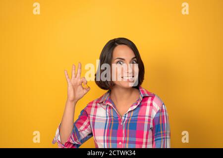 Die junge Frau zeigt ihren Fingern das Zeichen okay. Brunette lächelt und legt ihre Finger in den Ring auf einem gelben Hintergrund. Hochwertige Fotos Stockfoto