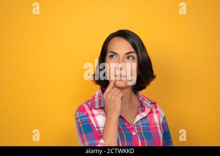 Brunette Frau macht einen brütenden Blick auf einem gelben Hintergrund im Studio. Frau legt ihren Finger auf ihre Lippen und schaut mit einer gewölbten Augenbraue nach oben Stockfoto