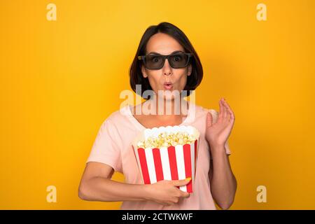 Frau in dunklen Gläsern hält Schachtel Popcorn. Junge Brünette mit einem überraschten Blick hob ihre Hand. Im Studio auf gelbem Hintergrund ausschneiden. Kino Stockfoto