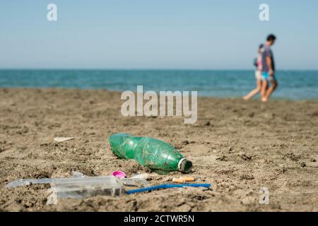 Grüne Plastikflasche, Wattestäbchen, Mütze, Schachtel und Zigaretten, die an einem Sandstrand mit Wanderern im Hintergrund hinterlassen wurden. Stockfoto